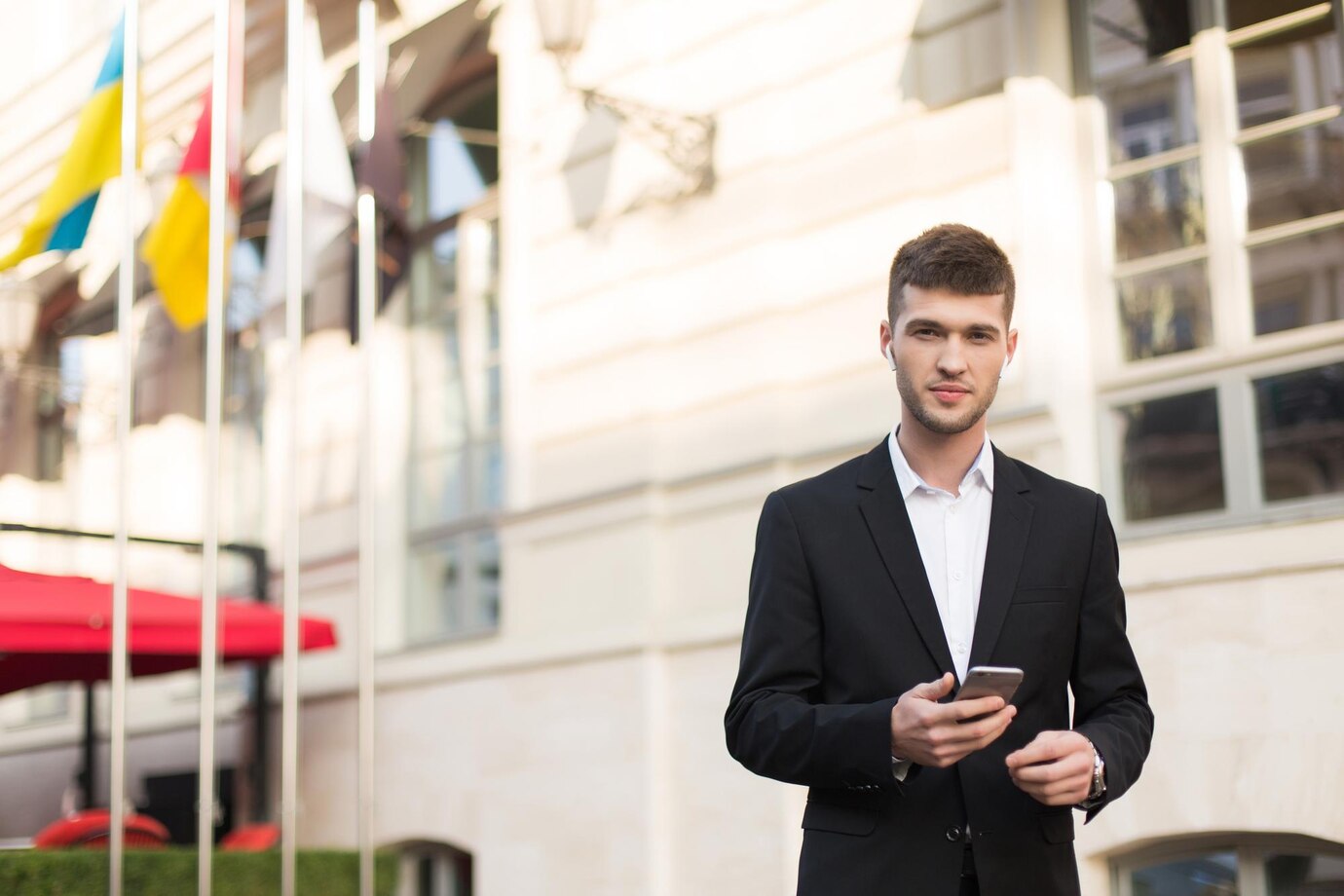 hombre guapo joven traje negro camisa blanca auriculares inalambricos sosteniendo telefono celular mano mientras mira sonadoramente camara al aire libre 574295 624